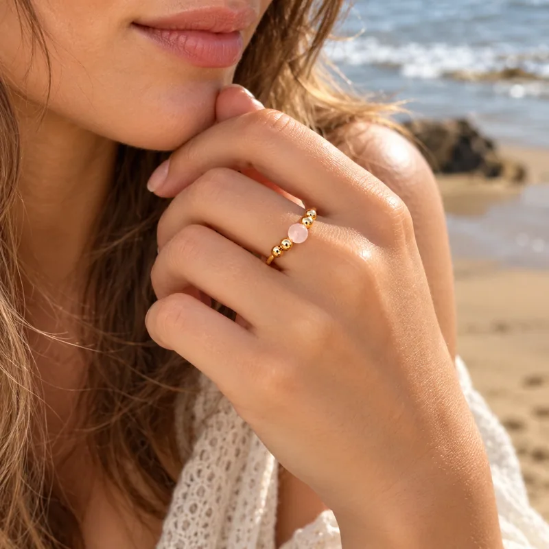 Femme portant la bague Valentina dorée à perle de quartz rose, main contre le menton sur la plage